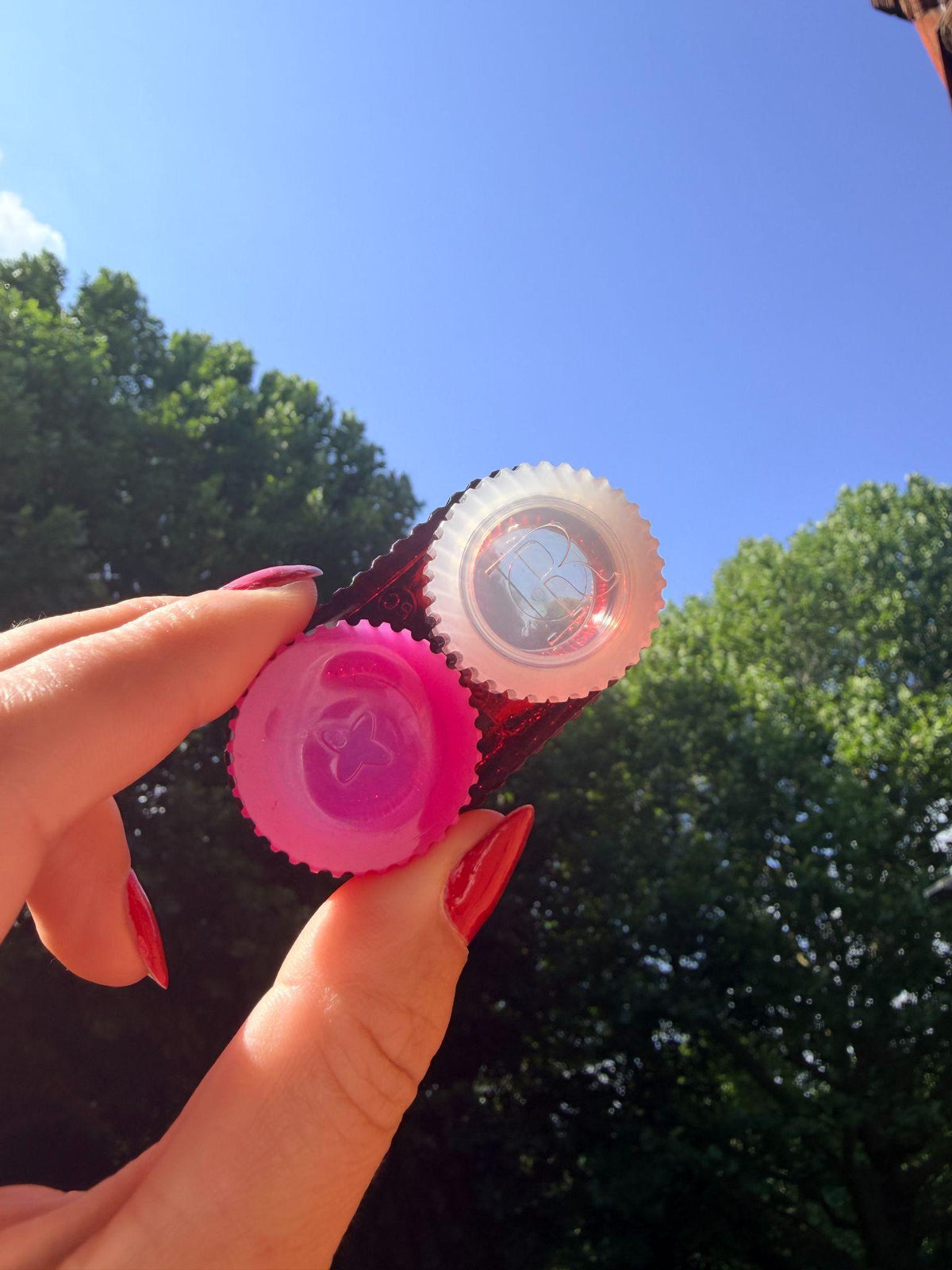 A hand with red manicured nails holds two small circular plastic pieces outdoors against a bright blue sky and green trees. One piece is bright pink, and the other is translucent white with a star-like pattern.